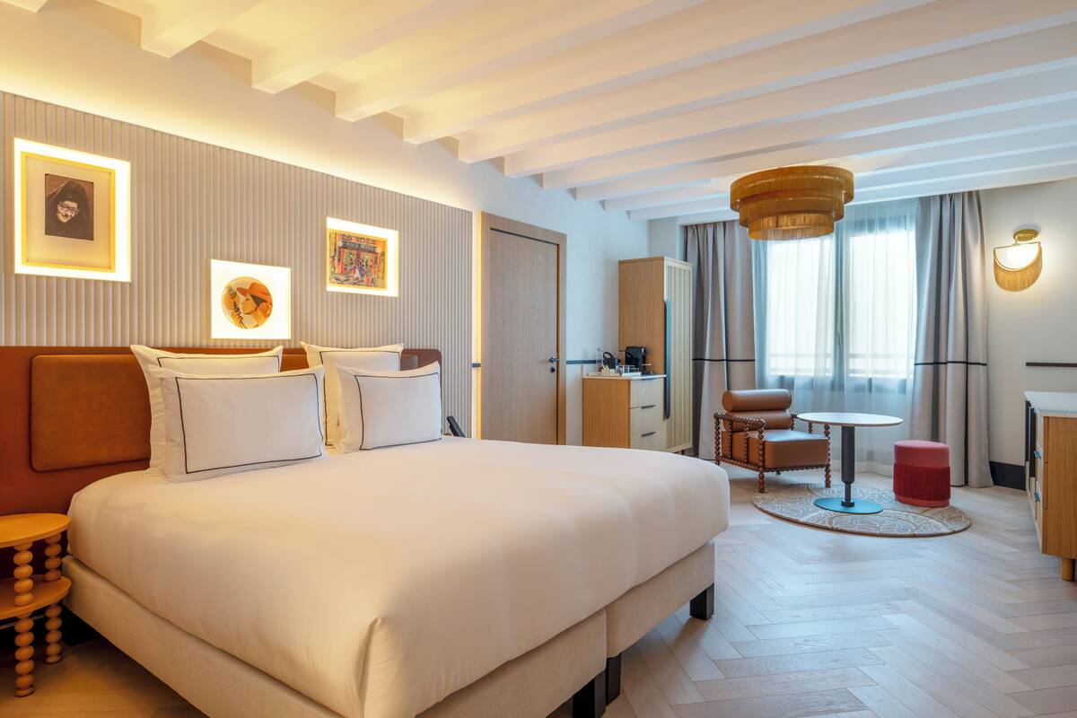 Ein Hotelzimmer mit einem großen Bett, einem Schreibtisch, einem Stuhl, einem kleinen Tisch, einem Kleiderschrank und einem Fenster mit Vorhängen.