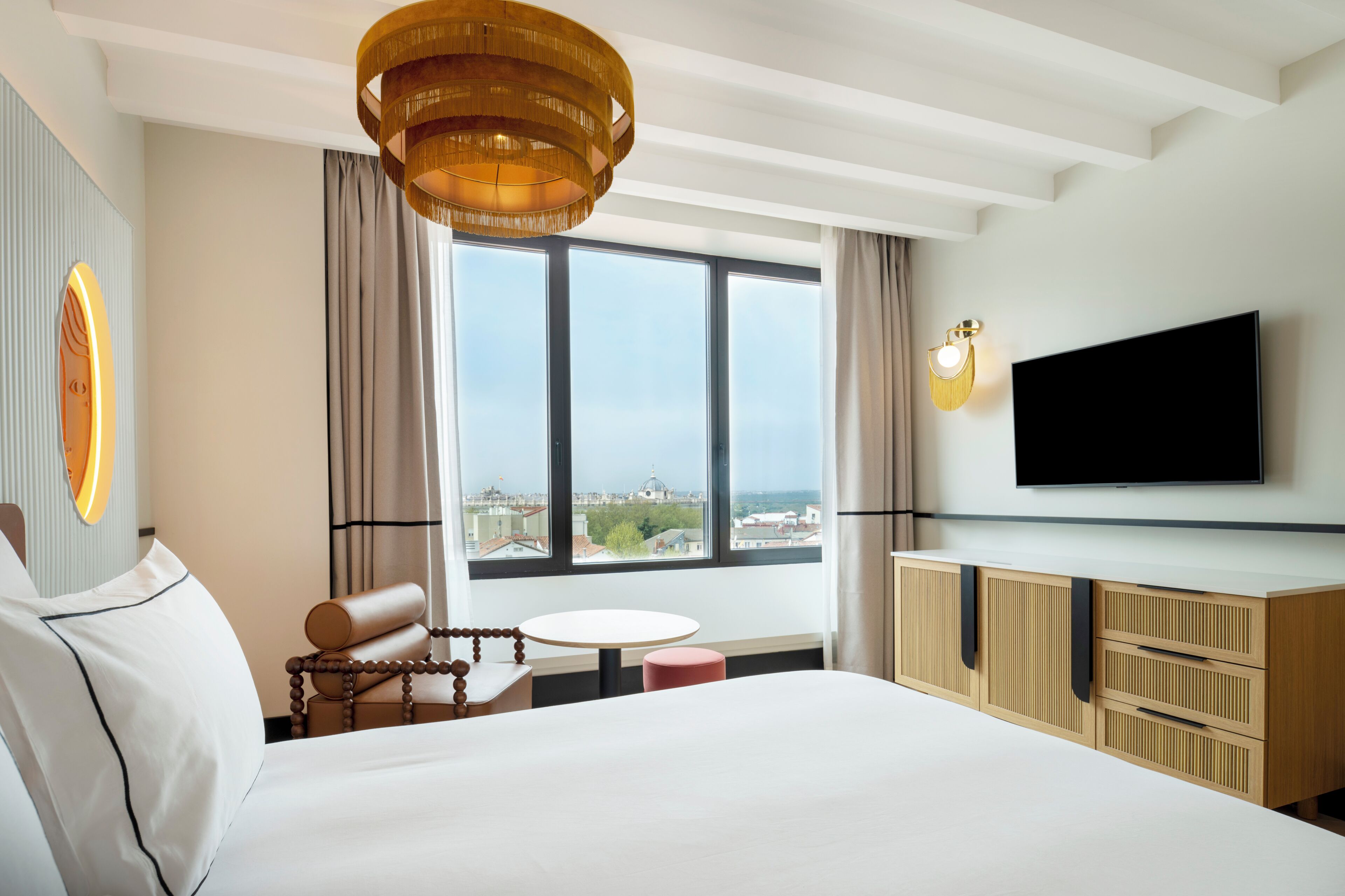 Habitación de hotel con una cama grande, televisión, una mesa de comedor y vistas de la ciudad a través de amplias ventanas.