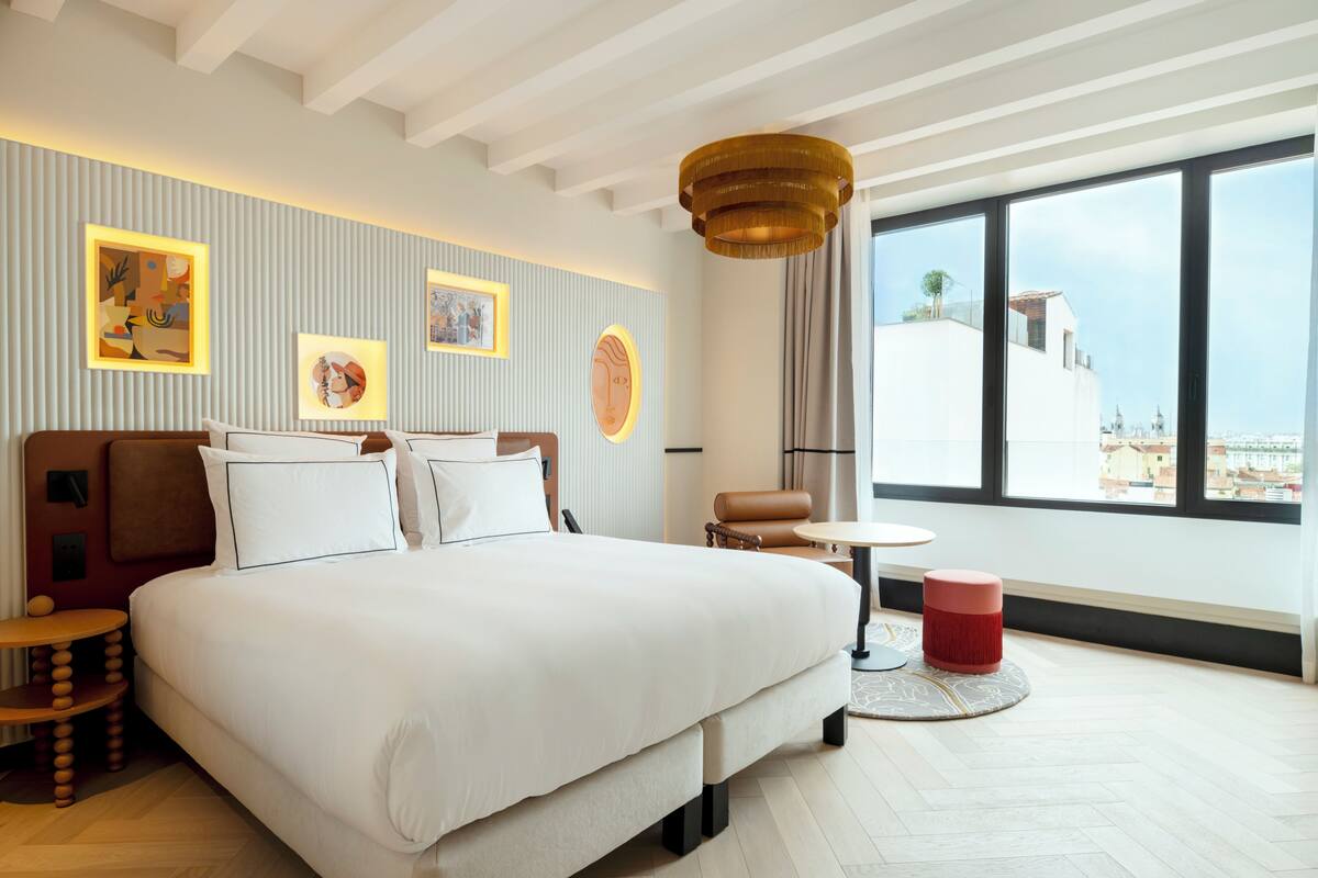 Ein modernes Hotelzimmer mit einem großen Bett, einem runden Tisch, einem roten Hocker und Kunstwerken an den Wänden.