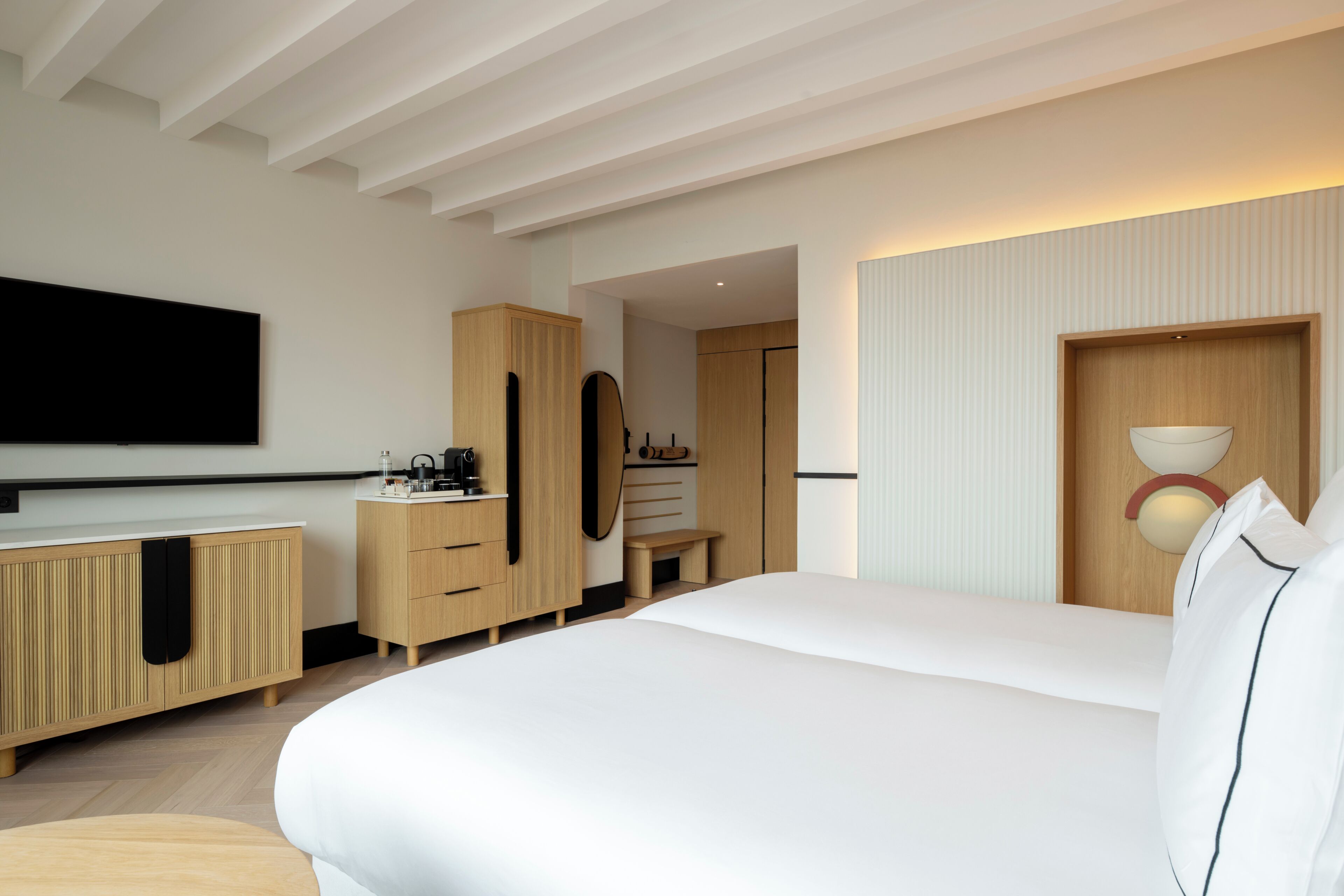 A modern hotel room with a large bed, wooden furniture, a flat-screen TV, and a wall-mounted light fixture.