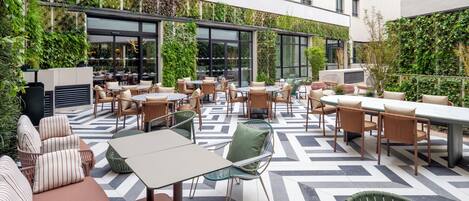 Un patio al aire libre con mesas y sillas, rodeado de vegetación y un suelo con diseño.