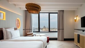 A hotel room with a large bed, a small round table, a sofa, a TV, and a view of a cityscape through large windows. - Urban Hive Madrid (Madrid)