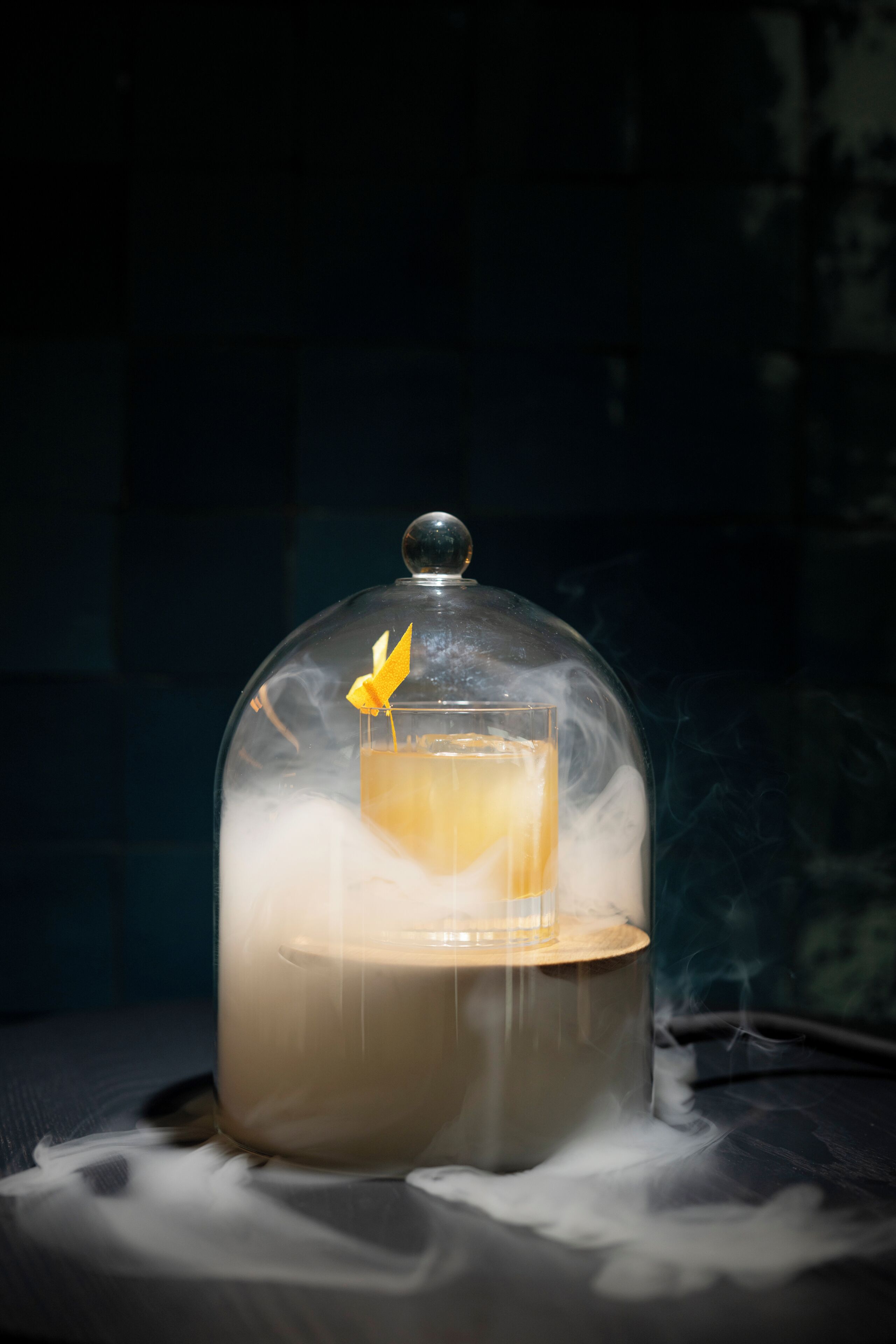Un vaso de líquido color ámbar con un adorno amarillo, cubierto por una campana de cristal con efectos de humo.