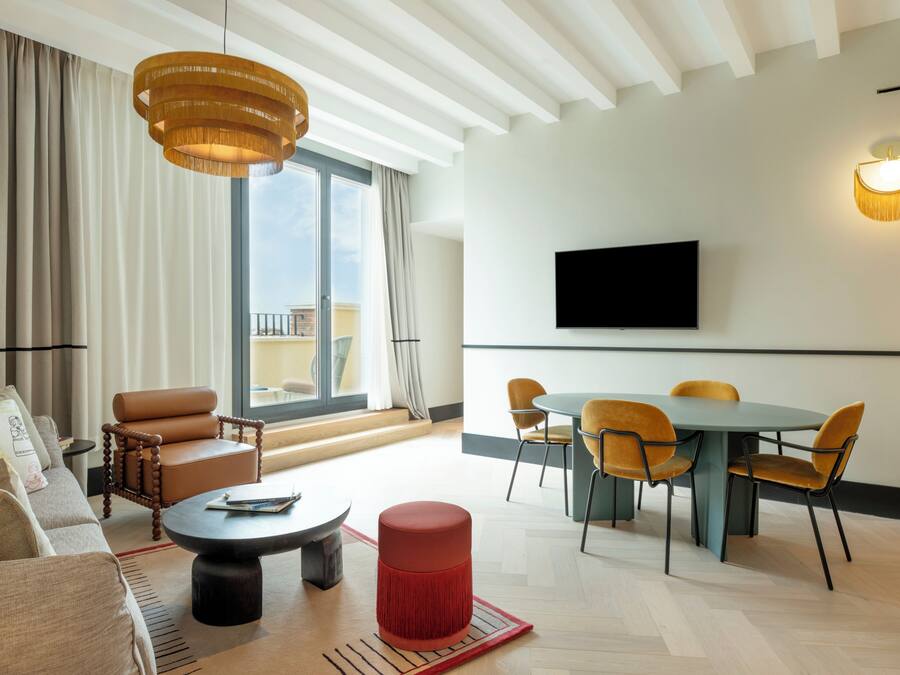 A modern hotel room with a large window, a dining area, a sofa, a red ottoman, and a flat-screen TV.