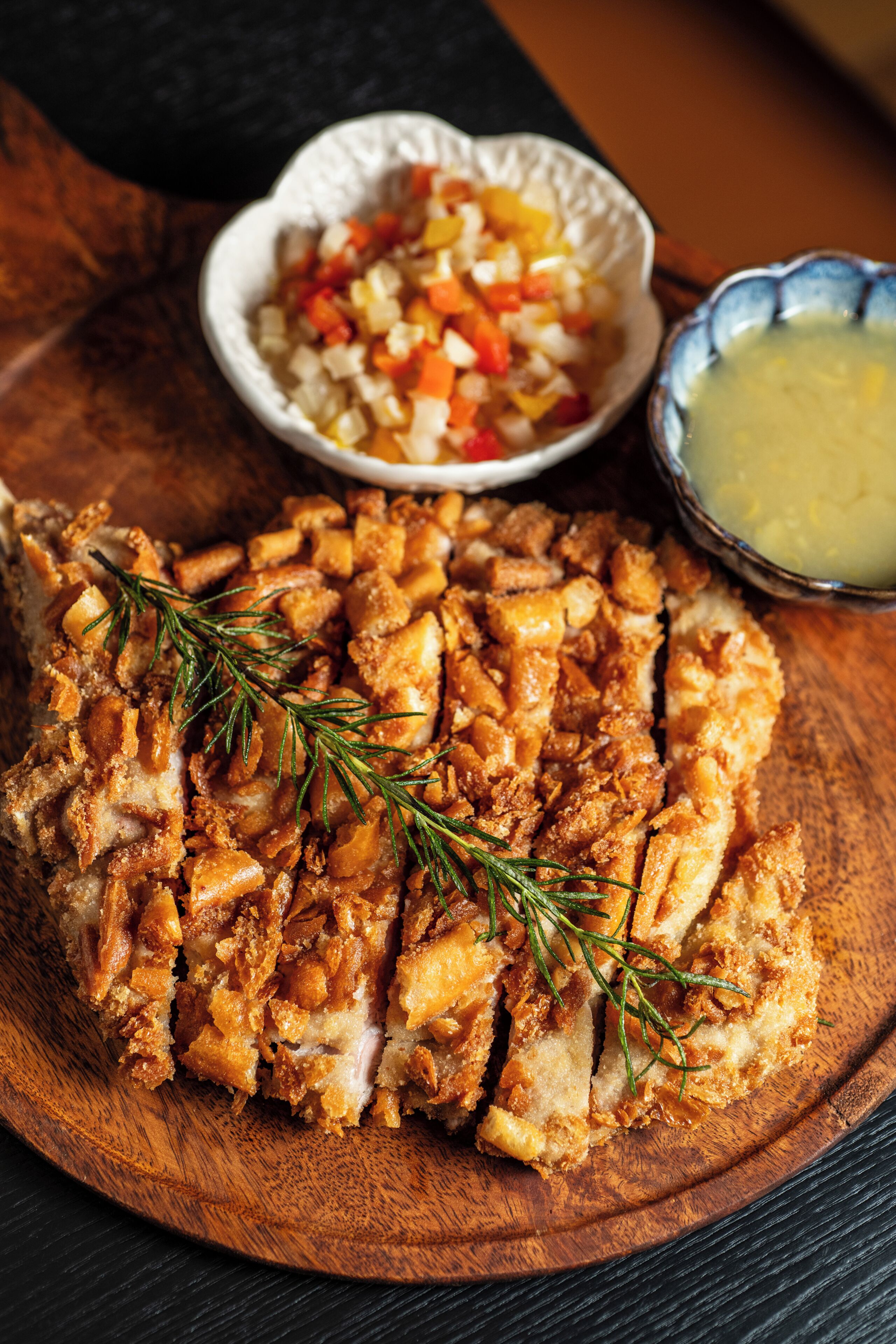 A wooden cutting board with sliced meat garnished with rosemary, a bowl of diced vegetables, and a small bowl of sauce.
