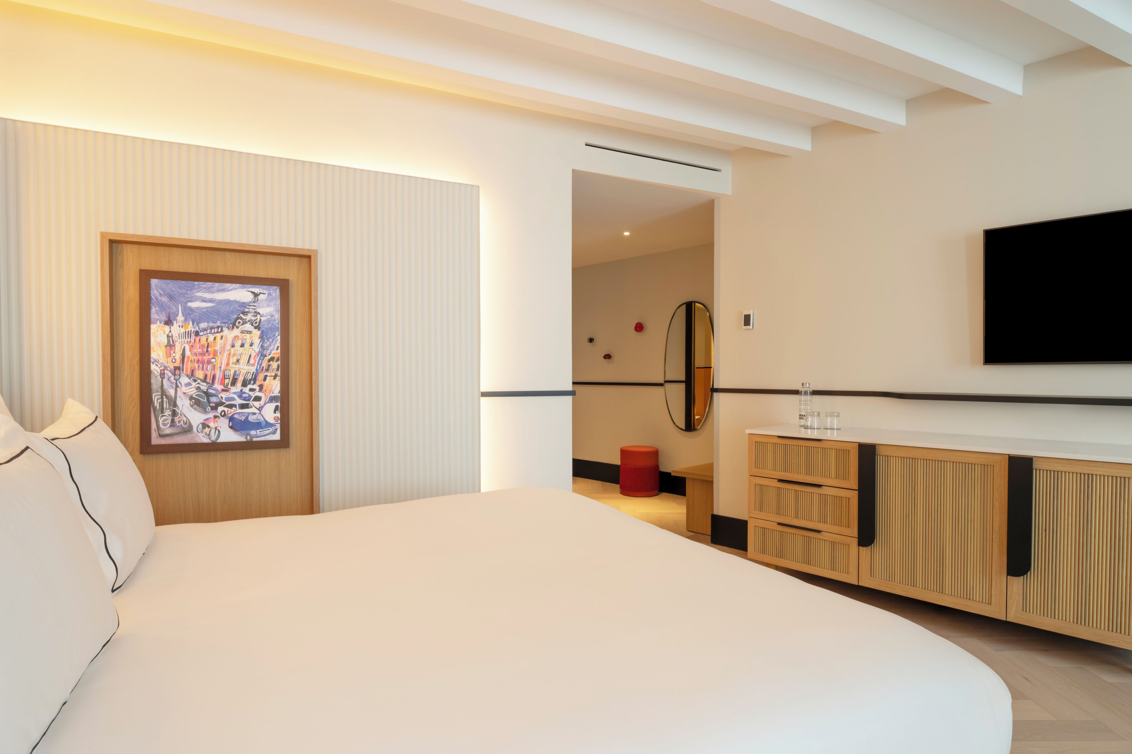 A modern hotel room with a large bed, a painting on the wall, a TV mounted on the wall, and a wooden dresser.