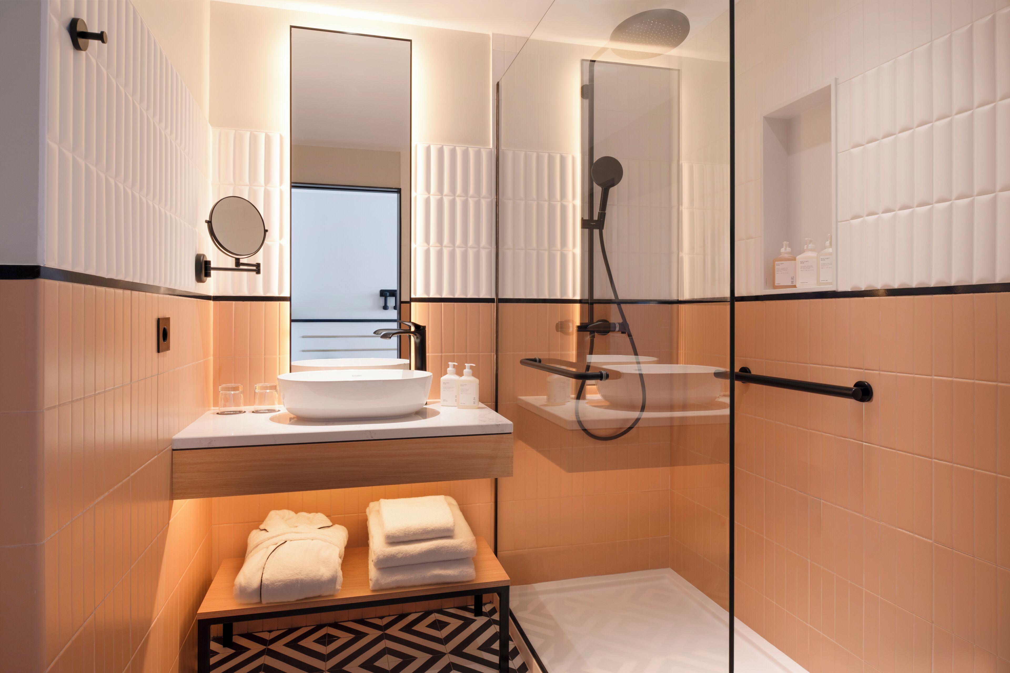 A modern bathroom with a glass-enclosed shower, a round mirror, a sink with a bowl, and a wooden bench with towels.