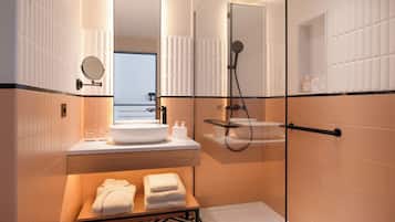 A modern bathroom with a glass-enclosed shower, a round mirror, a sink with a bowl, and a wooden bench with towels.