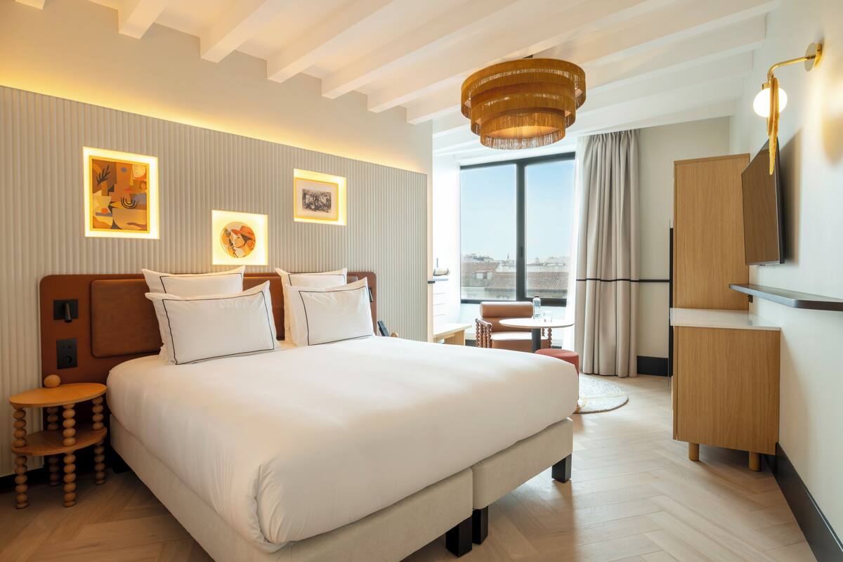 Ein modernes Hotelzimmer mit einem großen Bett, einem kleinen runden Tisch, einem Schreibtisch, einem Kleiderschrank und einem großen Fenster mit Blick auf die Stadt.