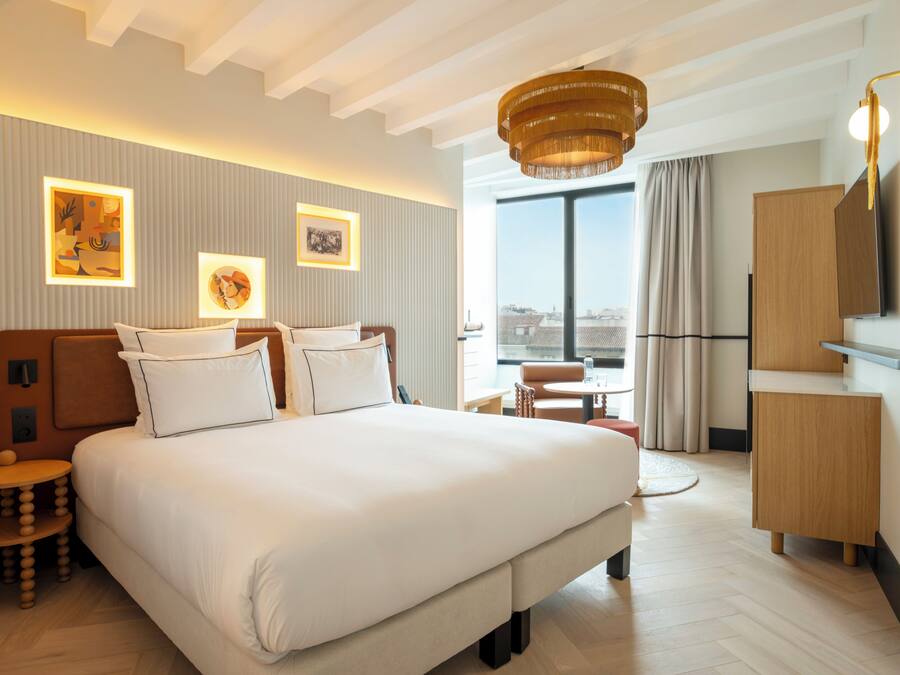 A modern hotel room with a large bed, a small round table, a desk, a wardrobe, and a large window offering a city view.