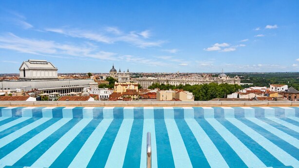 Ein Swimmingpool mit blauen und weißen Streifen, ein großes Gebäude mit Kuppel und eine Stadtlandschaft im Hintergrund.