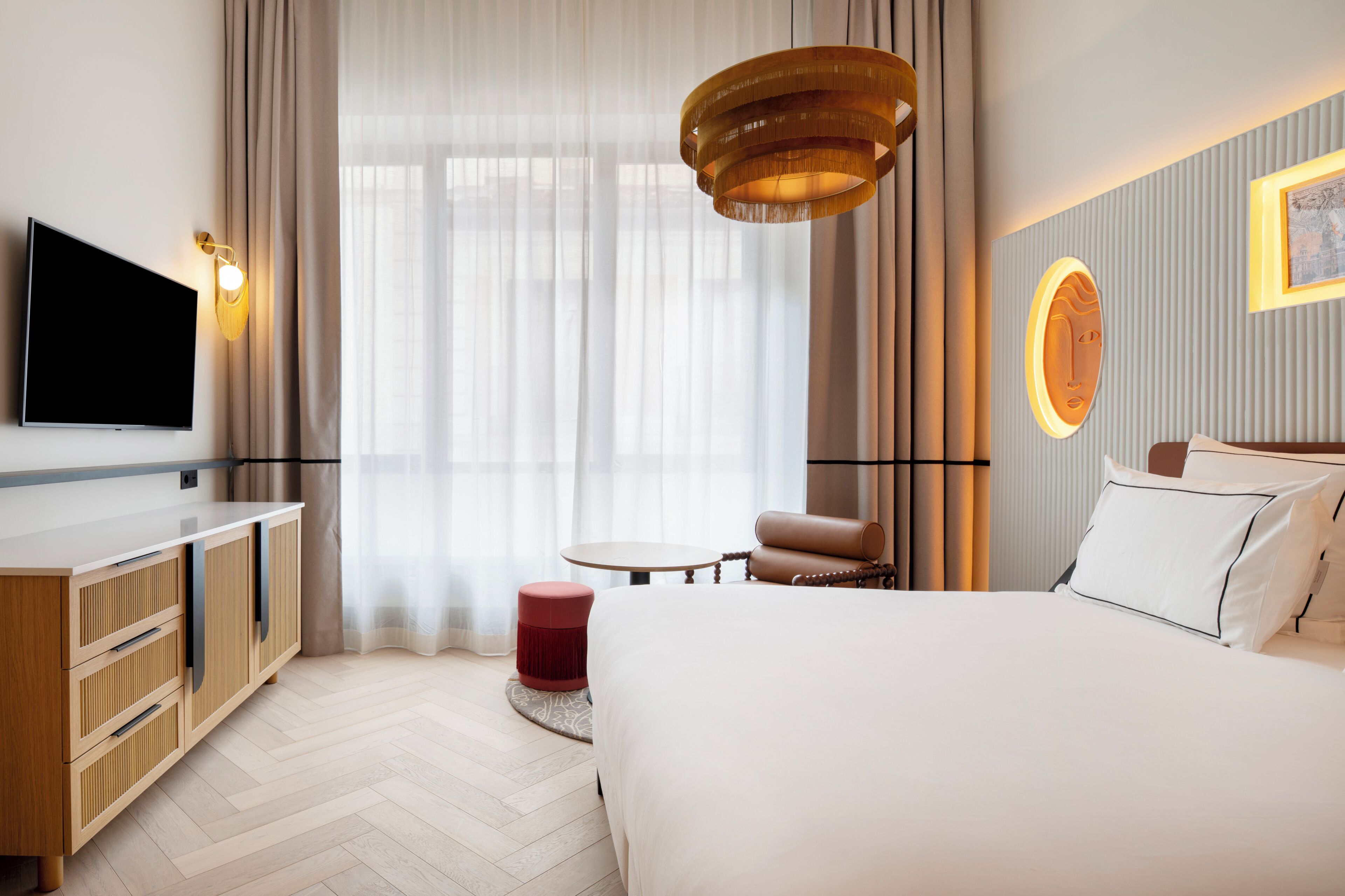 A hotel room with a bed, a TV, a round table, a red ottoman, and a wall-mounted light fixture.