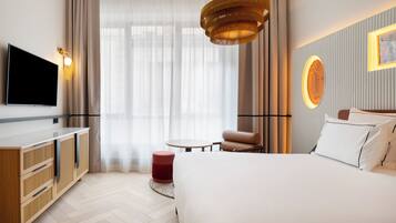 Habitación de hotel con cama, televisor, una mesa redonda, un reposapiés rojo y una lámpara de pared.