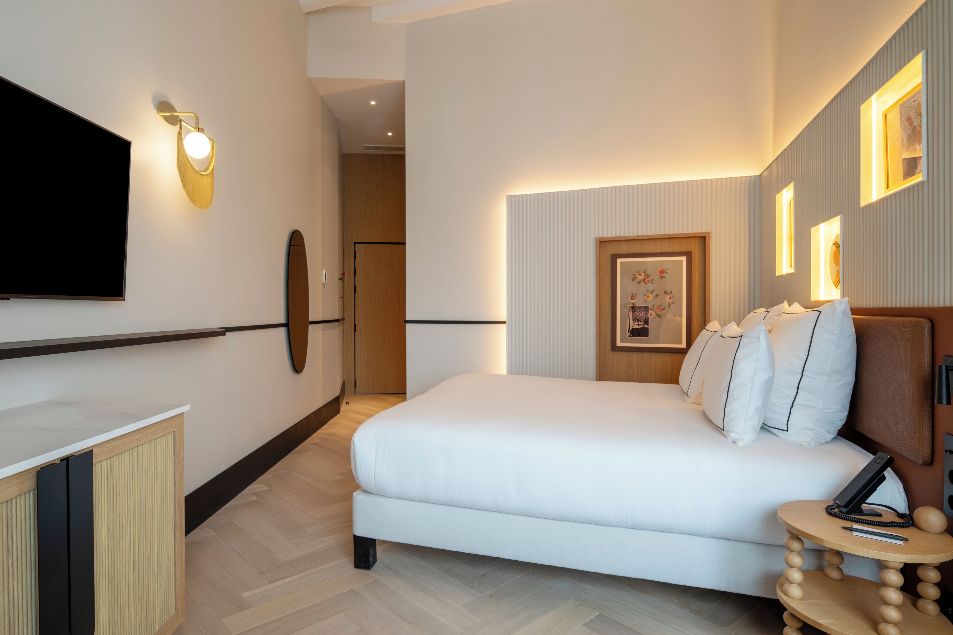 Een moderne hotelkamer met een groot bed, een flatscreen-televisie aan de muur, een klein rond tafeltje met telefoon en pen, en een ingelijste foto.