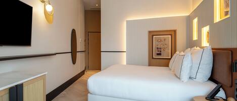 Una habitación de hotel moderna con una cama grande, un televisor de pantalla plana montado en la pared, una pequeña mesa redonda con un teléfono y un bolígrafo, y un cuadro enmarcado en la pared.