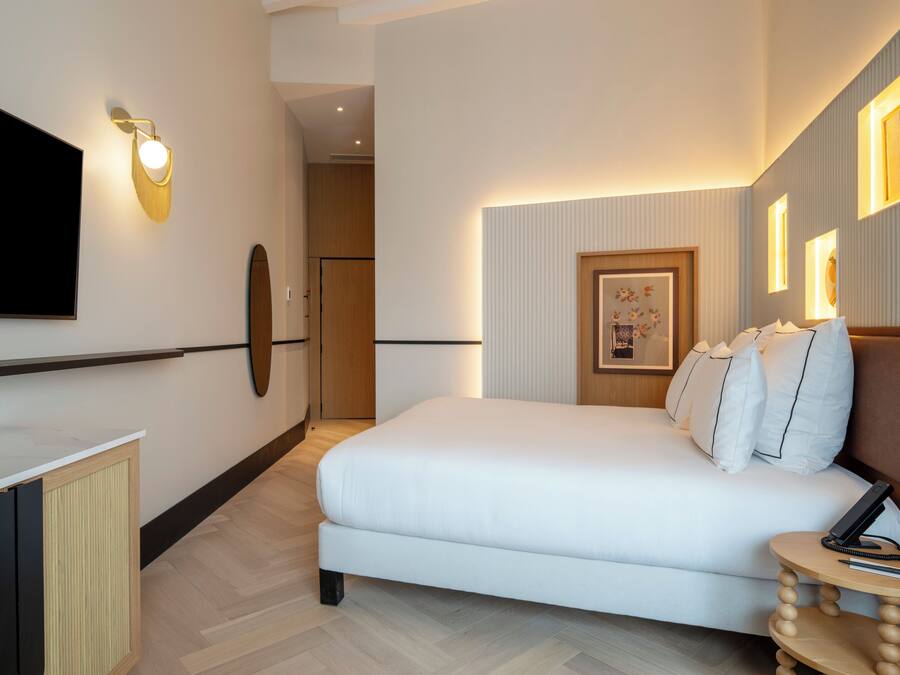 Una habitació d'hotel moderna amb un llit gran, un televisió de pantalla plana penjada a la pared, una taula rodona petita amb un telèfon i una ploma, i una fotografia emmarcada a la pared.