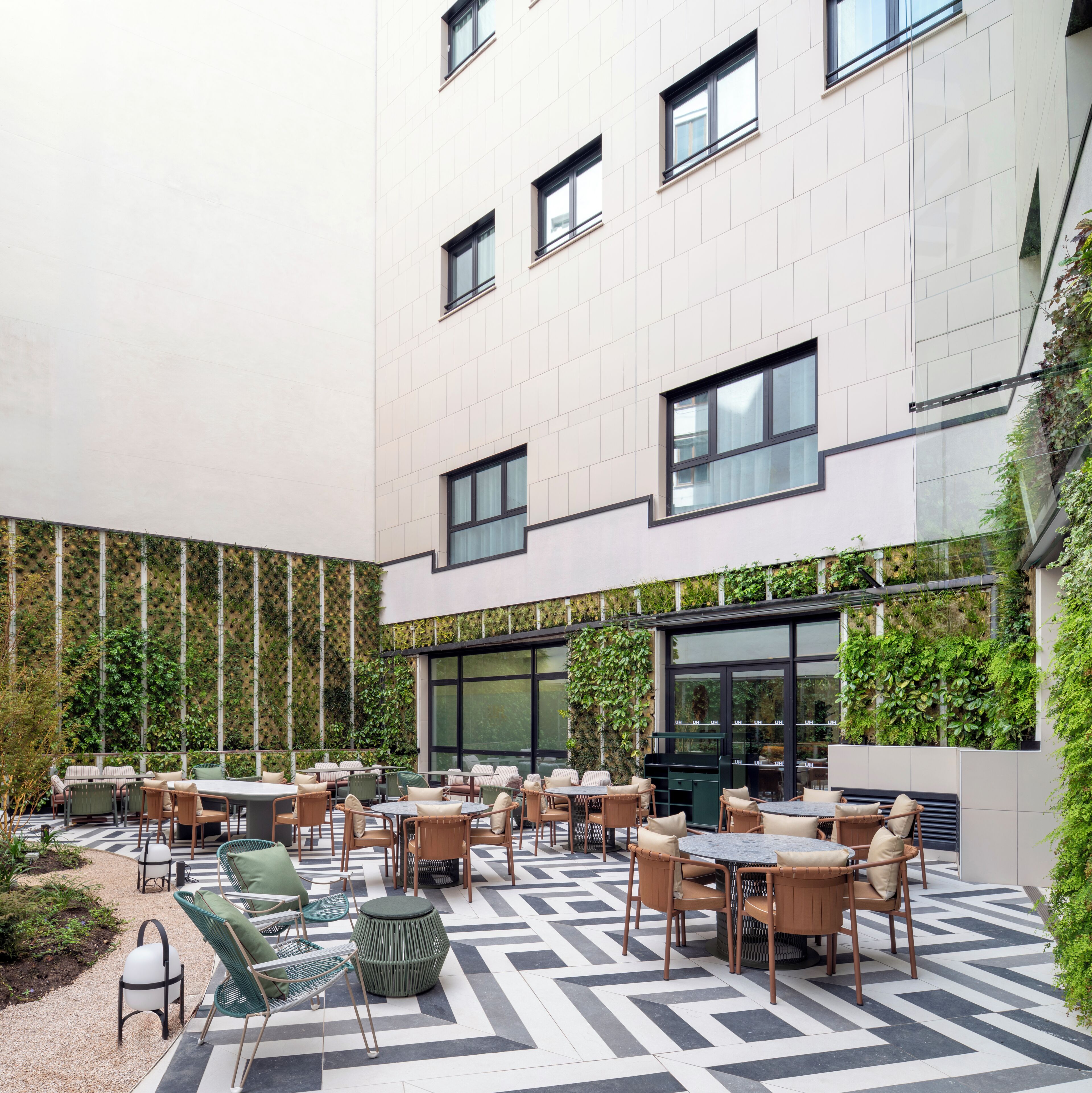 Un espace extérieur moderne aménagé avec plusieurs tables et chaises, entouré d’un bâtiment doté de grandes fenêtres et d’un jardin vertical verdoyant.
