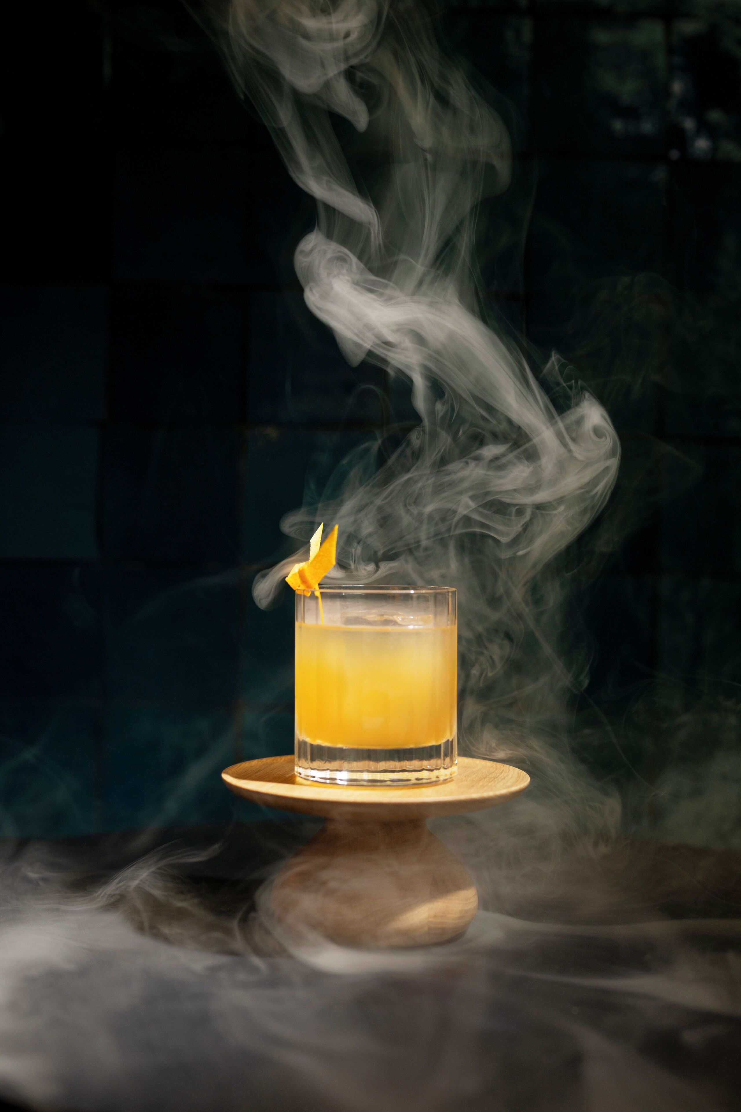 A glass of amber liquid on a wooden coaster with smoke rising from it.