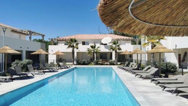 A swimming pool with lounge chairs and umbrellas, surrounded by buildings with a tiled roof. - Apartment T3 Marine de Bravone feet in the water (Linguizzetta)