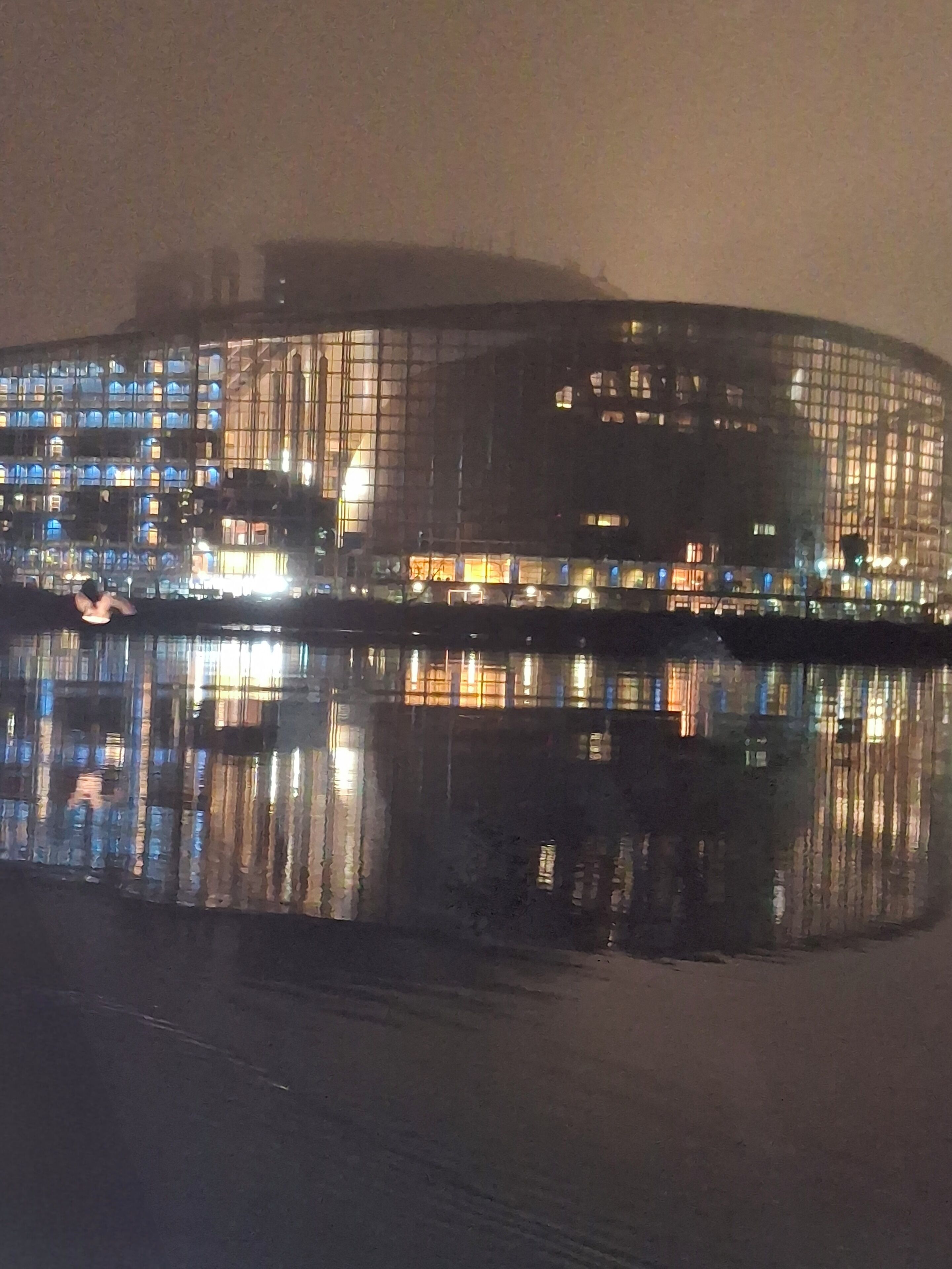 Un grand bâtiment moderne à la façade incurvée, dont le reflet se mire dans l’eau devant.