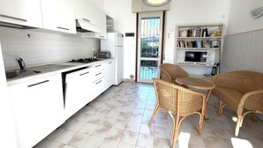Una cocina moderna con armarios blancos, una estufa a gas y un horno empotrado. Hay una zona de comedor con sillas de mimbre y una mesa, y una estantería con libros y un televisor.