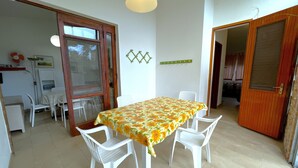 Een eetgedeelte met een tafel bedekt met een bloemen tafelkleed, omringd door witte plastic stoelen. Er is een houten deur en een witte muur met een decoratief object erboven.