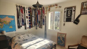 Ein Schlafzimmer mit einem Bett, das mit einem gemusterten Bettbezug bezogen ist, einem Schreibtisch mit Stuhl, einer Karte an der Wand und verschiedenen Sport-Schals, die über dem Bett hängen.