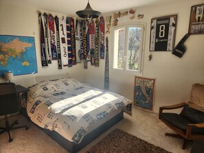 Ein Schlafzimmer mit einem Bett, das mit einem gemusterten Bettbezug bezogen ist, einem Schreibtisch mit Stuhl, einer Karte an der Wand und verschiedenen Sport-Schals, die über dem Bett hängen.