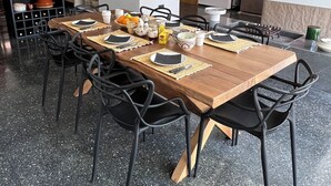 Een eethoek met een lange houten tafel gedekt voor zes personen, omringd door zwarte stoelen. Daarboven hangen twee grote geweven hanglampen.