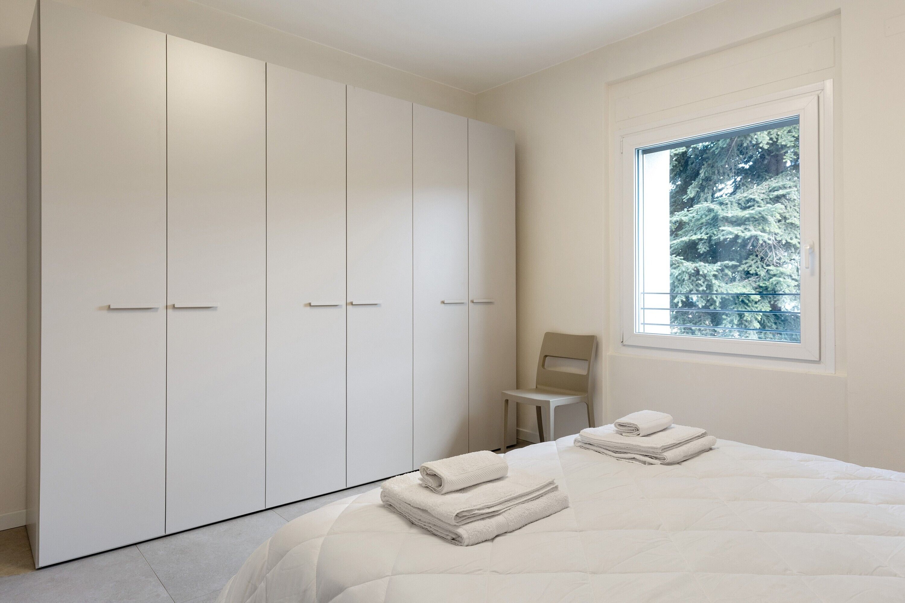 Une chambre dotée d’une grande armoire, d’un lit avec des serviettes soigneusement pliées, et d’une fenêtre donnant sur des arbres.