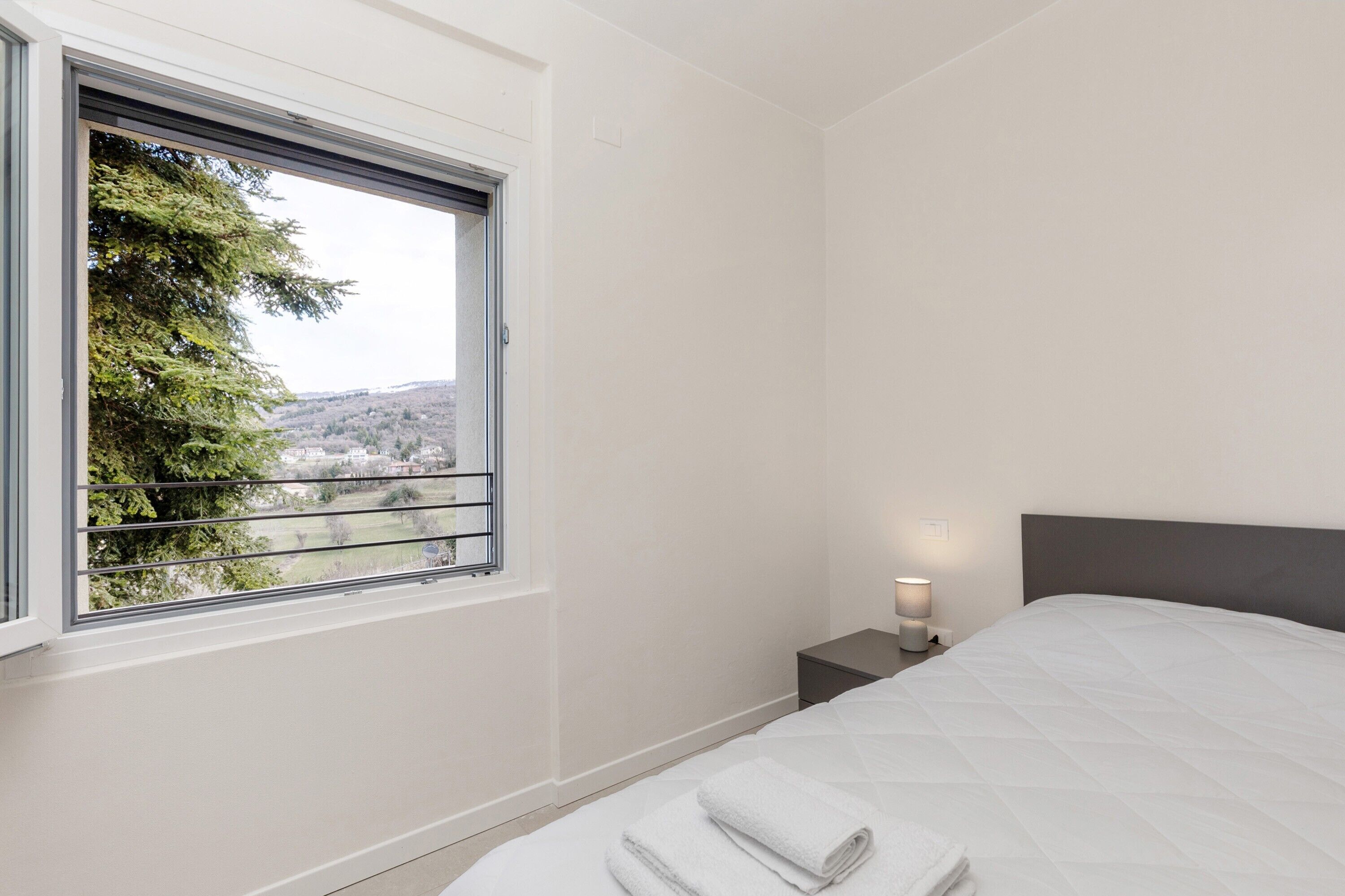 Un dormitorio con cama, mesita de noche, lámpara de noche y ventana con vistas a árboles y colinas.