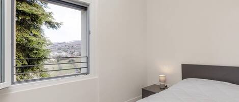 Un dormitorio con cama, mesita de noche, lámpara de noche y ventana con vistas a árboles y colinas.