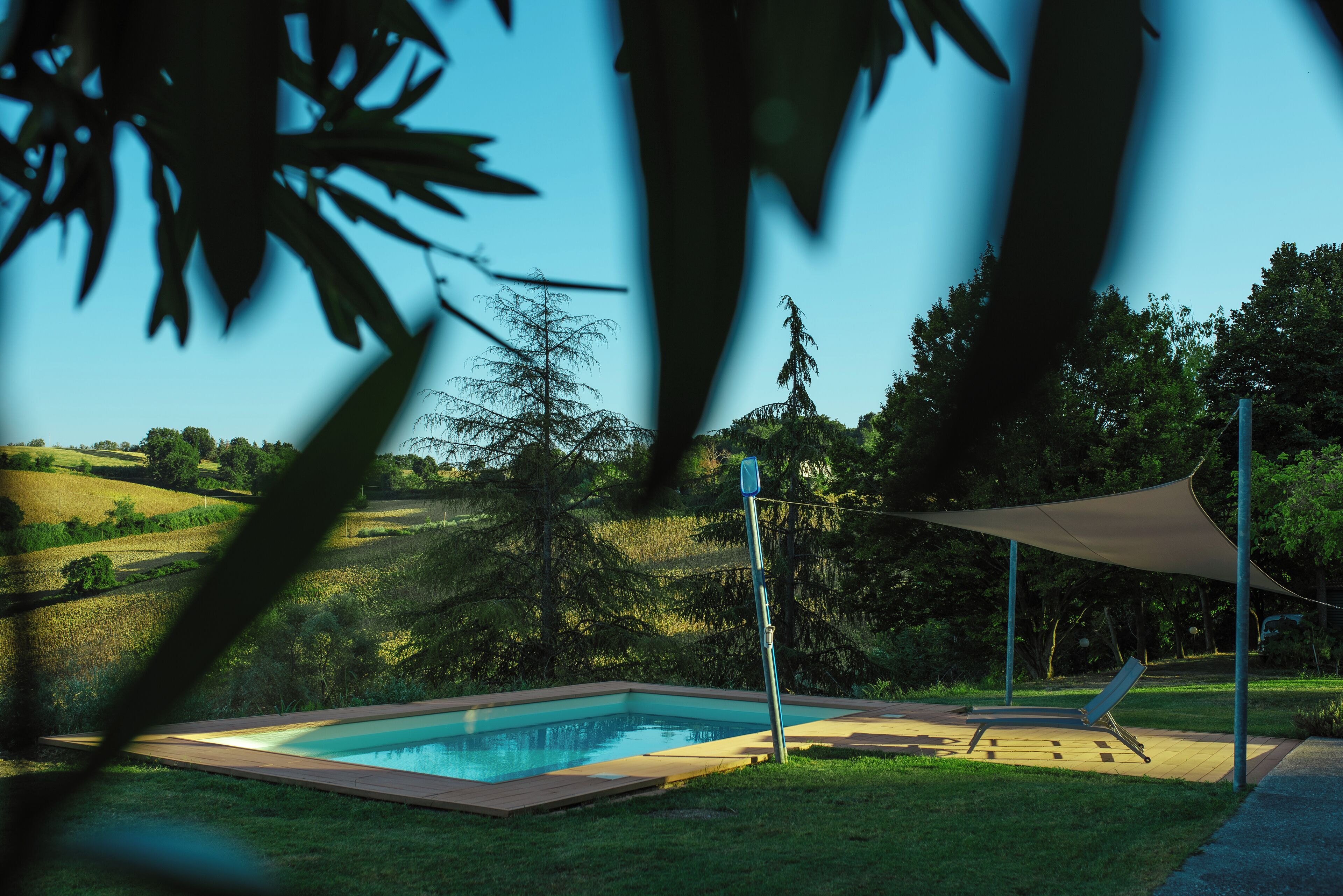 A swimming pool with a sun lounger and a shade sail, surrounded by greenery and hills.