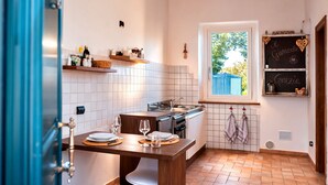 Une cuisine avec une table à manger dressée pour deux personnes, une fenêtre donnant sur un paysage verdoyant et un tableau noir sur lequel est écrit le mot « Grazie ».