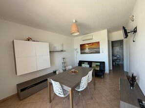 Ein Essbereich mit Tisch, Stühlen, einer Couch, einem an der Wand befestigten Regal, einem Gemälde und einem Flur, der zu einem Badezimmer führt.