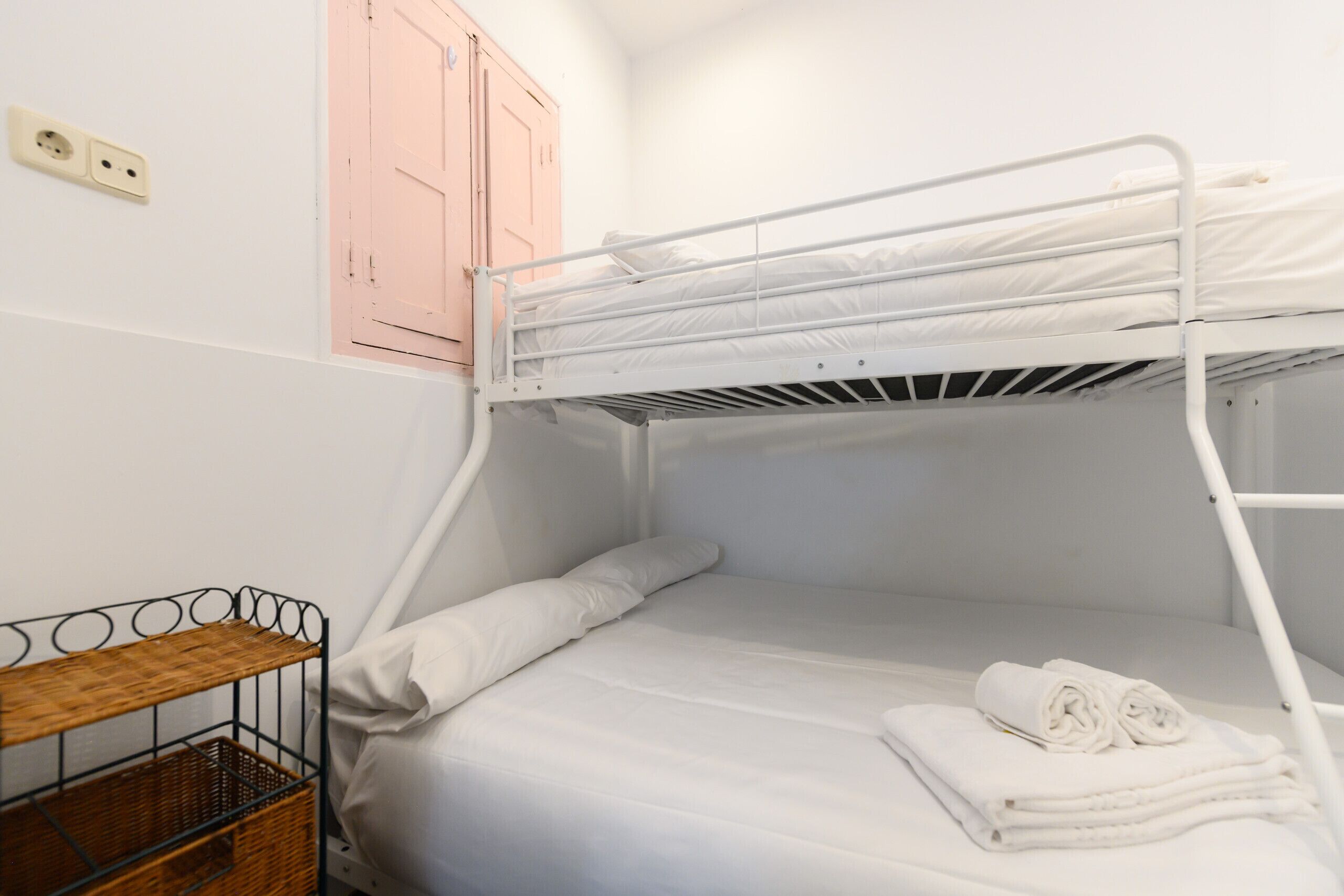 A bunk bed with white bedding and rolled towels, a wooden shelf, and a pink cabinet in a room with white walls.