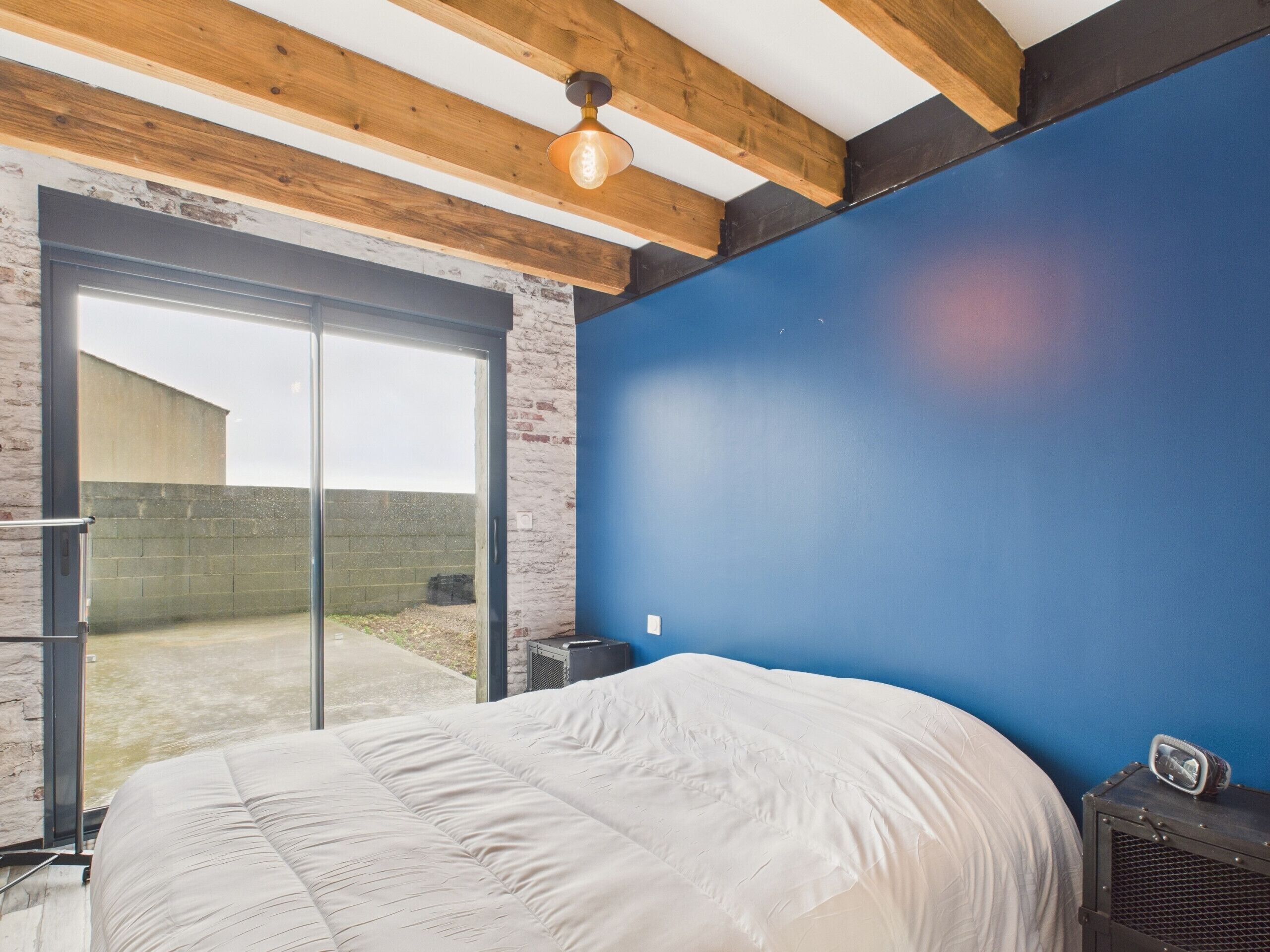 Een slaapkamer met een groot bed, een blauwe accentmuur en een glazen schuifdeur naar een buitenruimte.