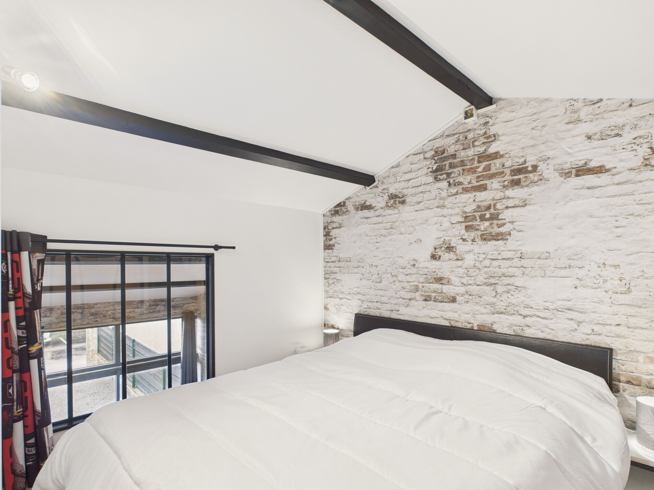A bedroom with a white bed, a brick wall, and a window with curtains.