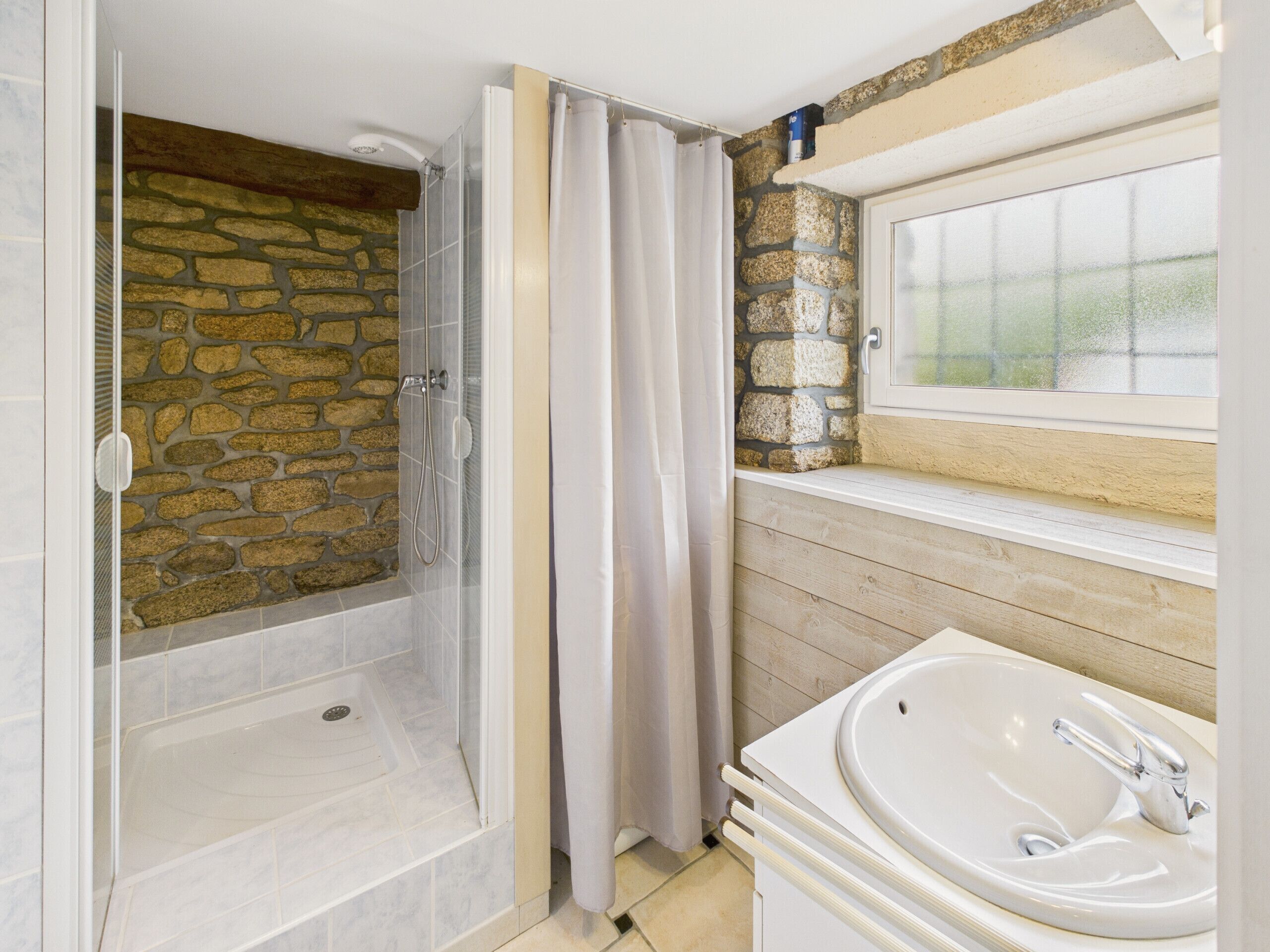 Un baño con pared de piedra, cabina de ducha de vidrio, bañera y ventana sobre el lavabo.