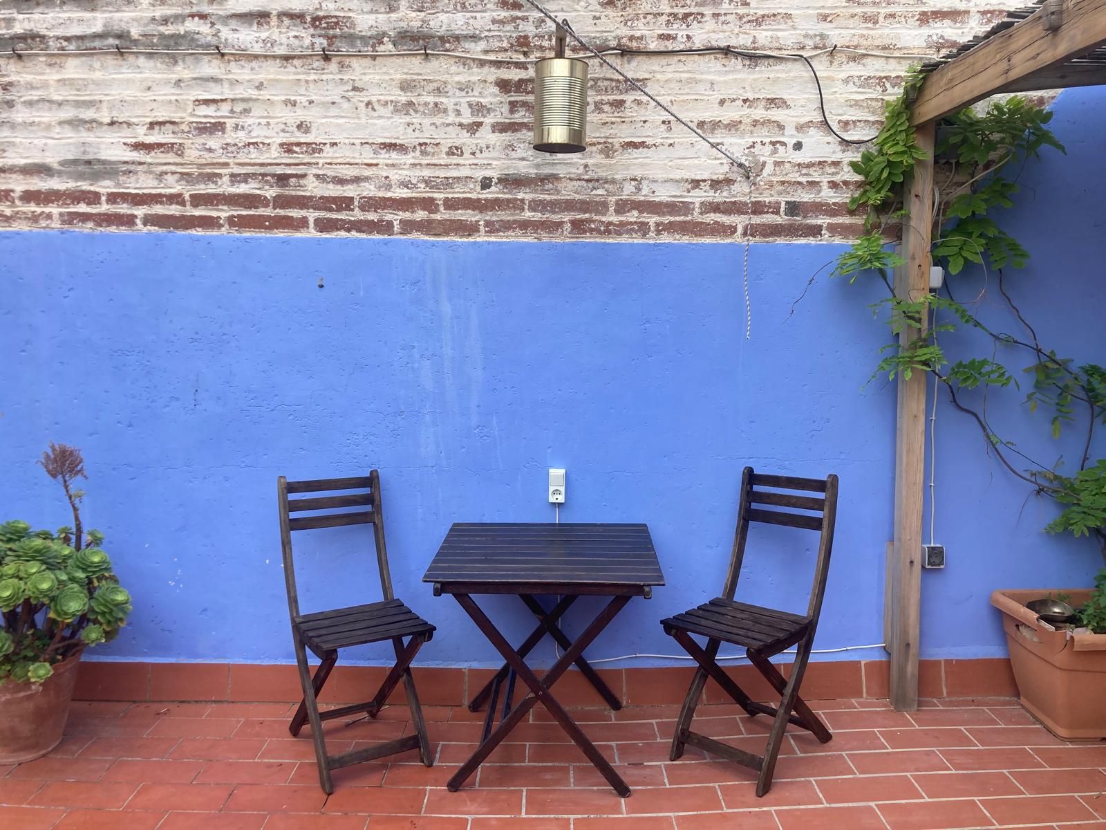 Um pátio com parede azul, duas cadeiras de madeira, uma mesa e um lustre suspenso.