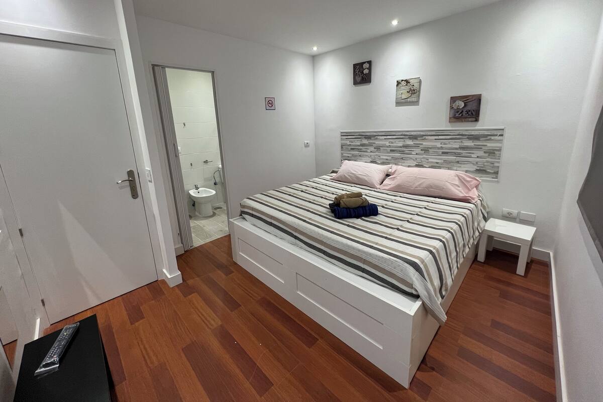 A modern bedroom with a large bed, a bedside table, a wardrobe, and a bathroom visible through an open door.