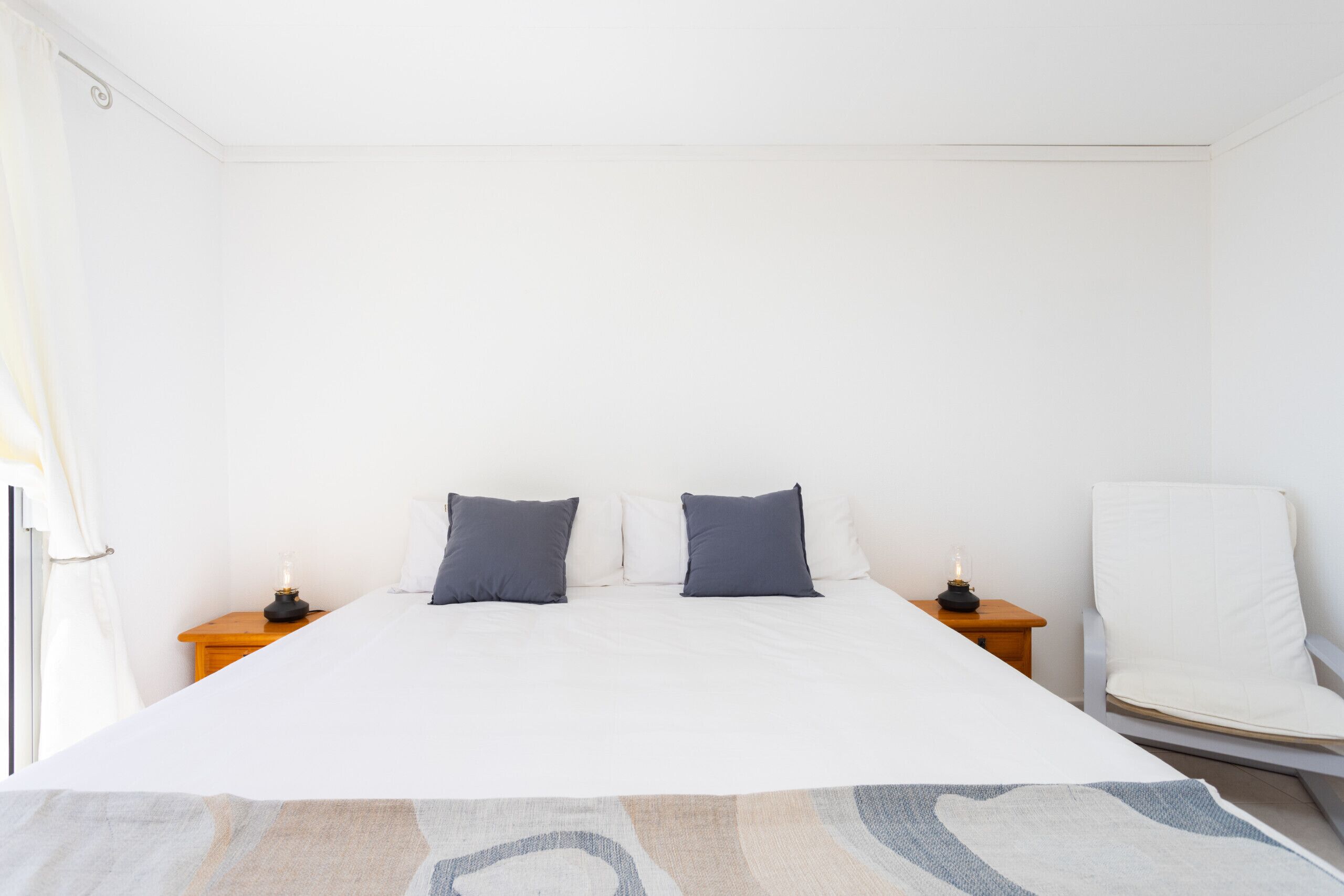 Una cama bien hecha con sábanas blancas y dos almohadones oscuros, flanqueada por dos mesitas de noche con lámparas, en una habitación sencilla con paredes blancas y una sola silla blanca.