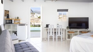 A modern, well-lit room with a sofa, a TV, a desk with a chair, and a view of a pool and buildings outside. - Oasis Tenerife studio D (Granadilla de Abona)