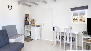 A modern living room with a grey sofa, white dining table with chairs, a small kitchen area with a microwave, and a window with a view of buildings. - Oasis Tenerife studio D (Granadilla de Abona)