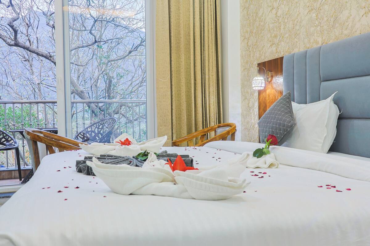 A hotel room with a bed, a view of trees through a window, and a balcony with chairs.