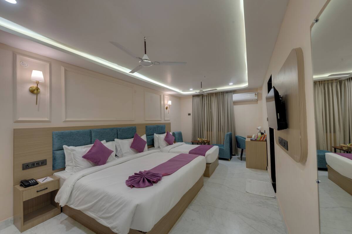 A hotel room with two beds, a ceiling fan, wall-mounted lamps, a TV, and a small table with a lamp.