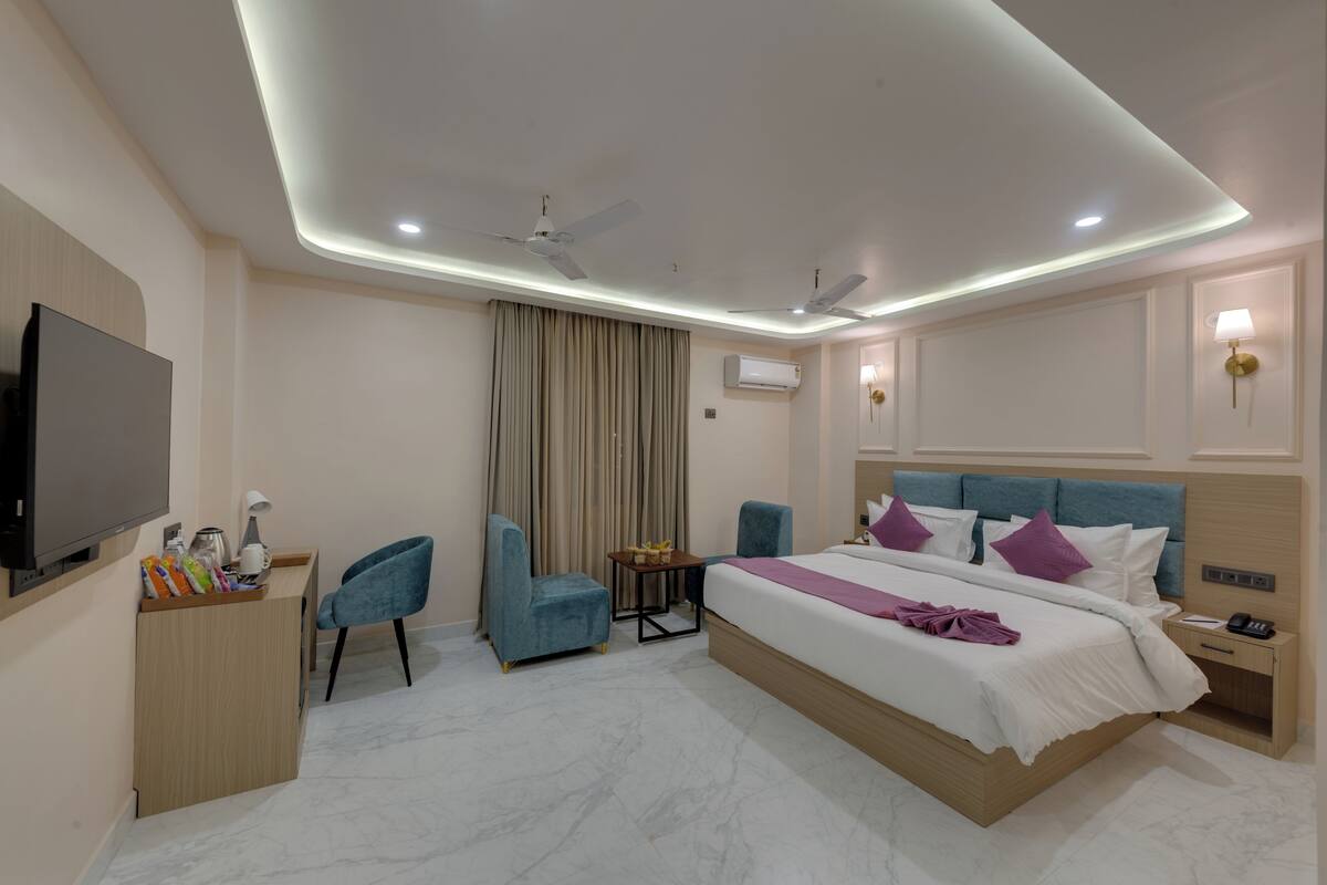 A modern hotel room with a large bed, two chairs, a desk with a flat-screen TV, and a ceiling fan.