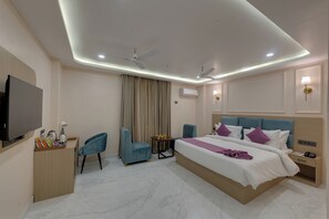 A modern hotel room with a large bed, two chairs, a desk with a flat-screen TV, and a ceiling fan.