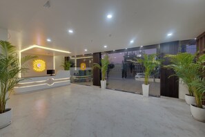 A modern hotel lobby with a reception desk, potted plants, and a glass partition.