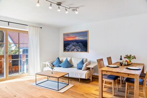 Una sala de estar moderna con sofá, mesa de comedor y sillas, un cuadro en la pared y vistas a través de un ventanal.